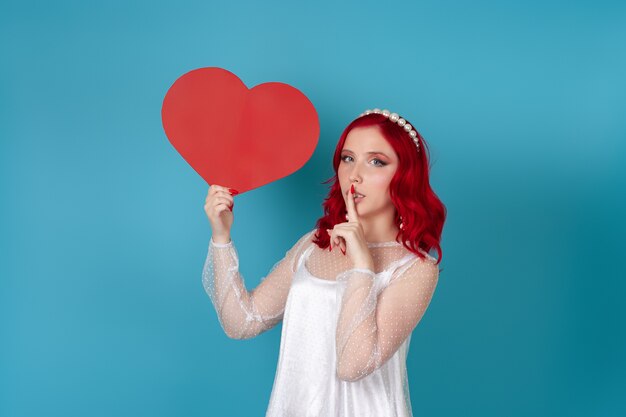 woman in a white dress and with red hair holds a large red paper heart and a finger at her mouth