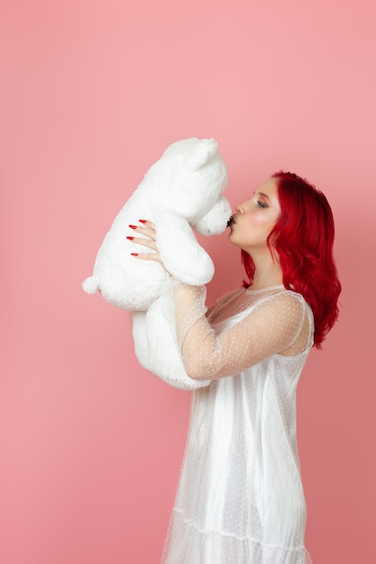 Woman in a white dress and with red hair kisses the nose of a large white teddy bear