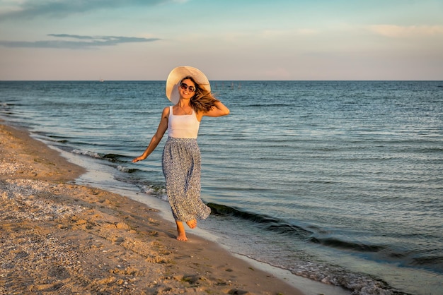 白い帽子をかぶった女性が夕日の浜辺を歩いている