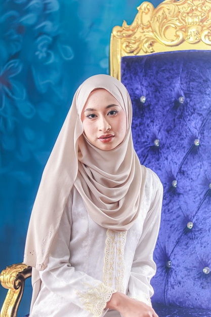 A woman in a white hijab sits in front of a blue velvet chair.