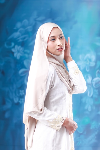 A woman in a white hijab stands in front of a blue background.