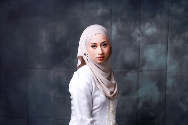 A woman in a white hijab stands in front of a grey wall.