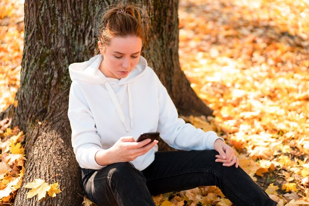 フード付きの白いパーカーセーターを着た女性が公園の地面に座って、携帯電話を手に持っています。かわいい女の子はコミュニケーションのためにスマートフォンを使用します女性のテキストメッセージメッセージコミュニケーションの概念