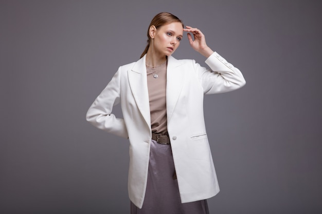 Женщина в белой куртке с длинной металлической серебряной юбкой позирует на сером фоне Студийный портрет