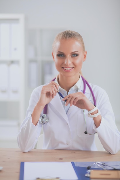 A woman in a white lab coat sits at a desk and smiles at the camera.