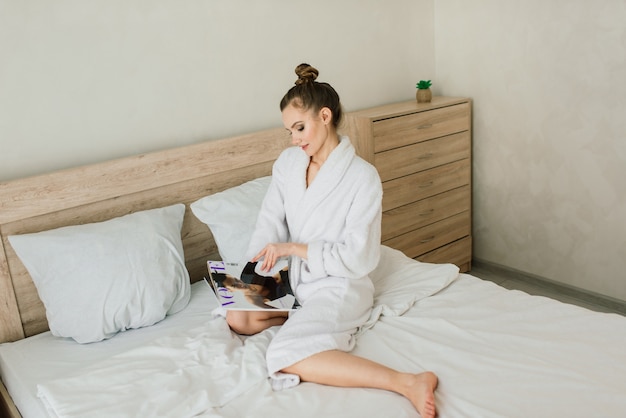 Woman in white robe stay near the window and on a bed in hotel room