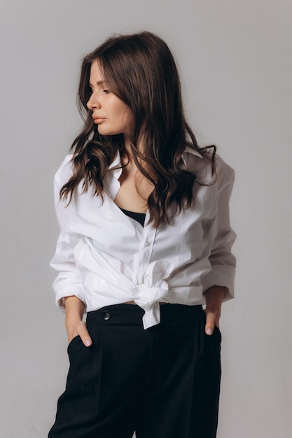 A woman in a white shirt and black pants stands in front of a grey background.