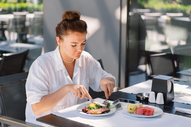 A woman in a white shirt eats lunch or breakfast outdoors in a cafe