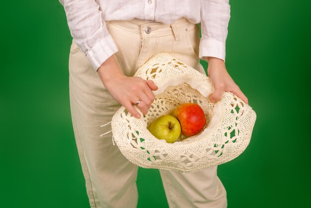 A woman in a white shirt holds apples in a hat on a green background. The concept of diet, healthy food, vegetarianism.