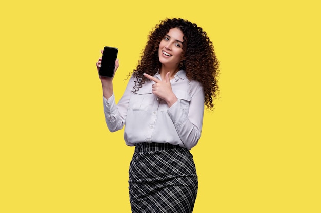 Woman in white shirt holds smartphone and points with index finger at blank screen. Businesswoman over yellow isolated background. Concept of online casino