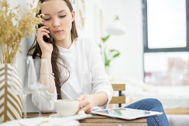 A woman in a white shirt is drinking coffee in a cafe A female office worker at a lunch break solves a business problem by phone Midlevel manager