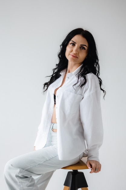 A woman in a white shirt and jeans sits on a stool.