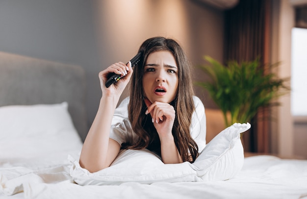 Woman in white silk pajamas watches tv in the bedroom on the bed. female model with a tv remote control in her hands lying on the bed. morning or evening in the bedroom with a female model.