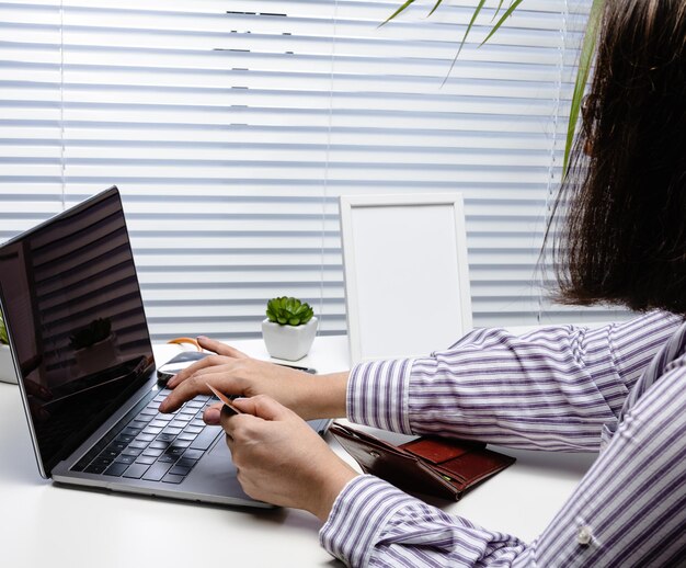 Woman in a white striped shirt makes purchases online with a laptop, a credit card in hand. A woman sitting at a white table