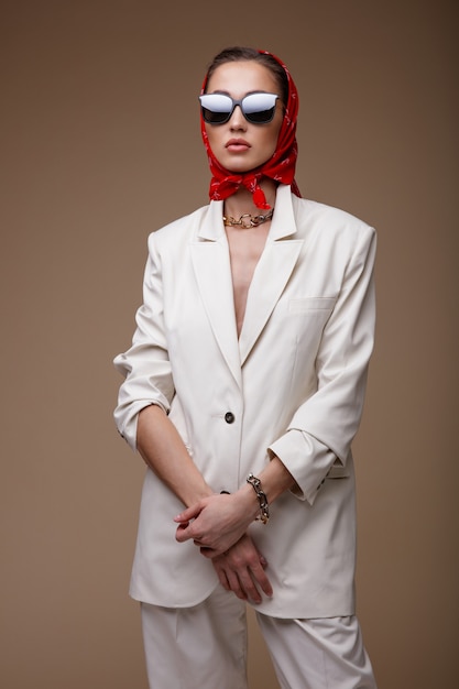 베이지색 배경에 흰색 정장 재킷 바지 신발 빨간색 스카프 선글라스 액세서리를 입은 여성