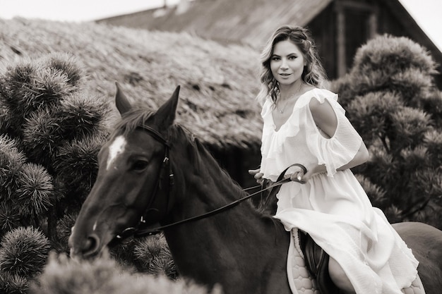 A woman in a white sundress riding a horse near a farm black and white photo