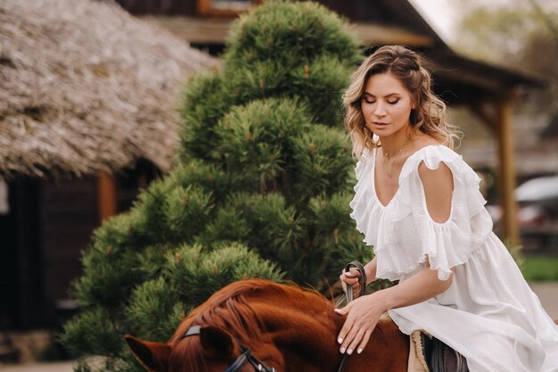 A woman in a white sundress riding a horse near a farm