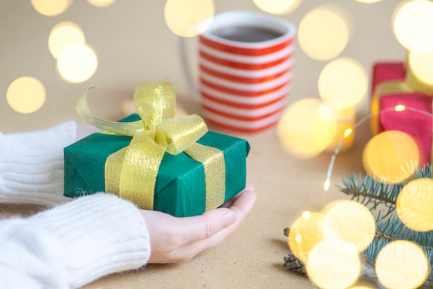 A woman in a white sweater holds a Christmas present on a wooden table background. Atmospheric photo with bokeh lights. Selective focus. Concept of gifts for Christmas and New Year.