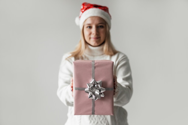 Donna con un maglione bianco e un cappello rosso da babbo natale che tiene in mano un regalo rosa su uno sfondo grigio