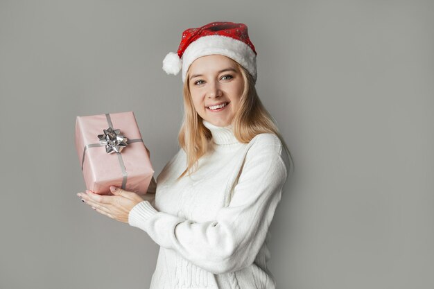 Woman in a white sweater and a red santa hat holding a pink gift in her hands on a gray background