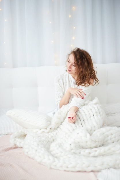 Woman in white sweater seating on bed. Magical morning in Christmas bedroom. Decorated Christmas tree. Christmas, winter holidays concept.