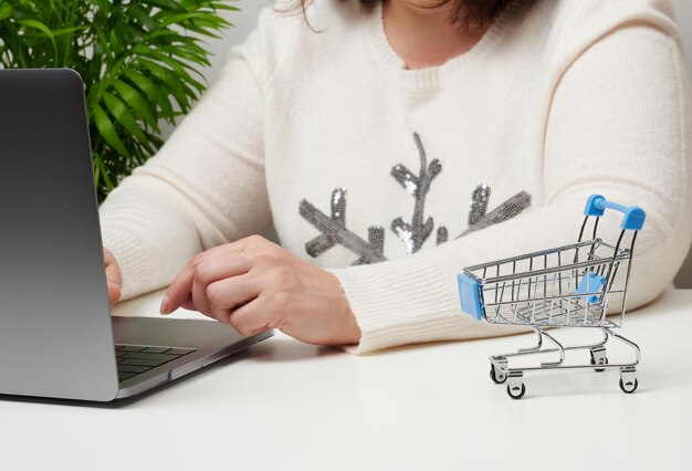 A woman in a white sweater sits and works at a laptop. Empty miniature shopping carts on white desk. Seasonal sale, online shopping