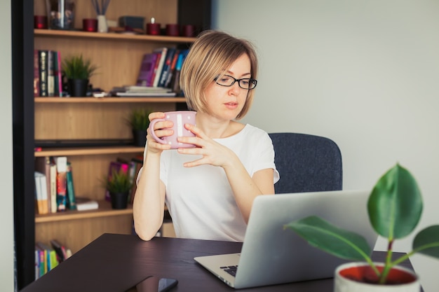Donna in maglietta bianca che tiene una tazza, lavorando da casa su un computer portatile