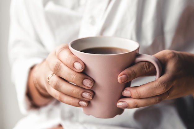 Woman in a white t-shirt holds morning coffee in a pink ceramic cup. Manicure. Front view