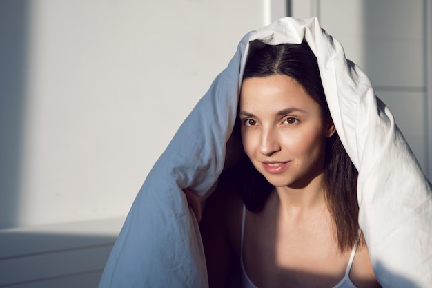 Woman in a white t-shirt is lying on the bed and a pillow is under the blanket in the room