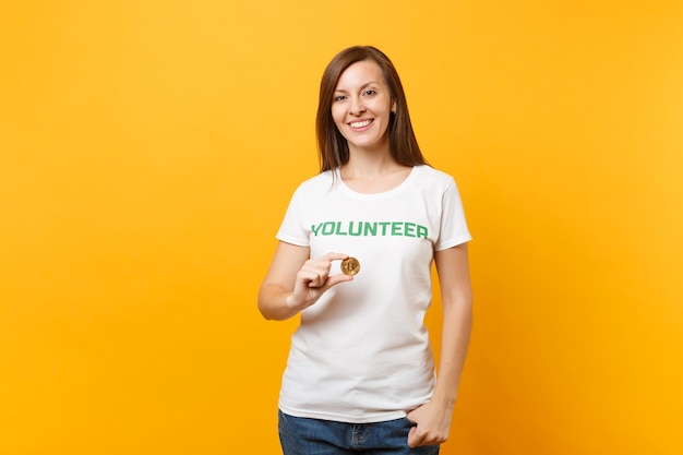 Woman in white t-shirt with written inscription green title volunteer hold bitcoin, metal coin future currency isolated on yellow background. Voluntary free assistance help, charity grace work concept