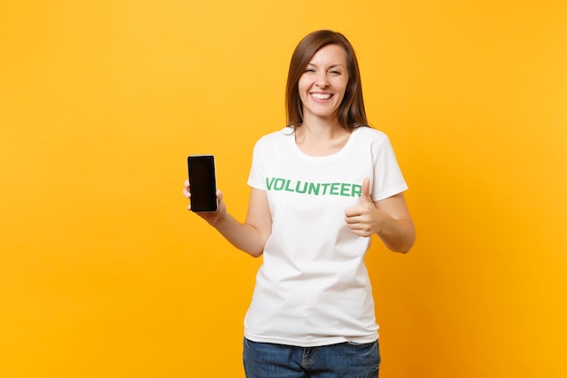 白いTシャツに書かれた碑文の緑のタイトルのボランティアの女性は、黄色の背景で隔離の空白の空の画面で携帯電話を保持します。自主的な無料支援支援、チャリティーグレイスワークコンセプト。