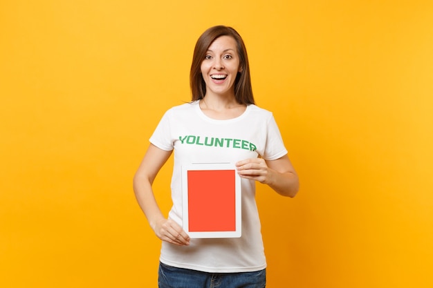 Woman in white t-shirt written inscription green title volunteer hold tablet pc computer, blank empty screen isolated on yellow background. Voluntary free assistance help, charity grace work concept.