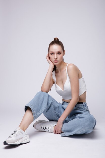 Woman in white top sneakers blue denim jeans on white background Studio Shot Model is sitting