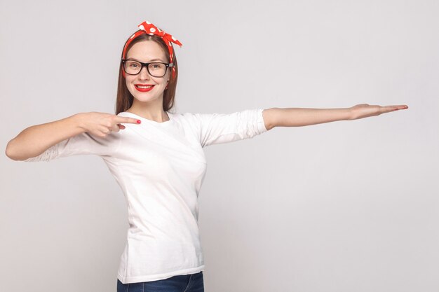 Woman in white tshirt with glasses red lips and head band pointing and showing copy space