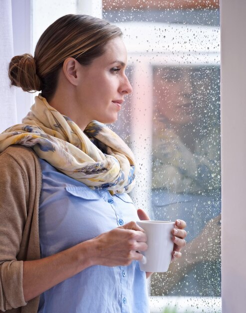 Woman at window thinking with tea and reflection life and future in morning routine raindrop and wellness Insight memory and mindfulness with warm beverage coffee break and relax at home