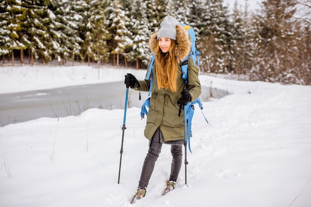 Woman in winter clothes hiking with backpack and tracking sticks at the snowy forest near the frozen lake