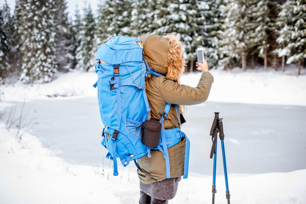 Woman in winter clothes with backpack and tracking sticks enjoying beautiful landscape view on the snowy forest and frozen lake