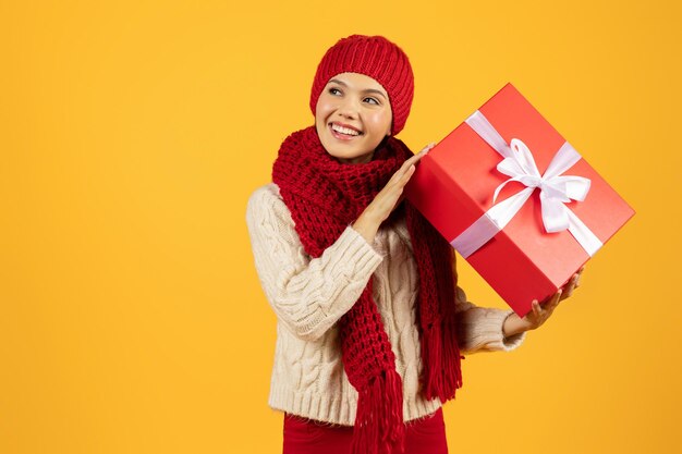 Woman in winter knitwear holds red gift box yellow background