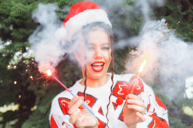 サンタ クロースの帽子と赤い唇を持つ冬のセーターを着た女性が、クリスマス ツリー新年クリスマスの背景に線香花火を保持します