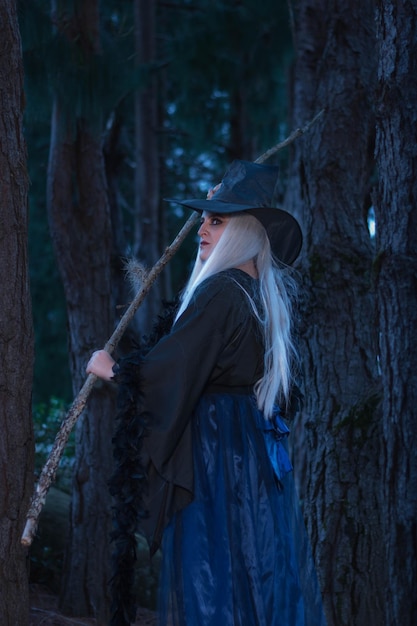 Woman in a witch costume in the forest at twilight holding a wooden stick