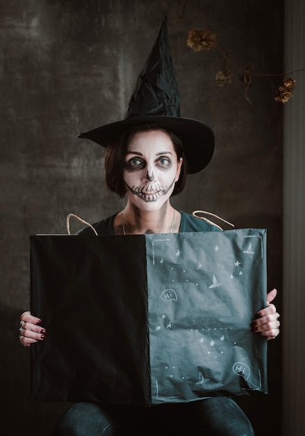 Woman in a witch costume and skeleton makeup opens a black spell book for a witch on Halloween.