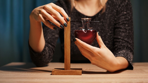 The woman witch prepares a potion. Red love potion in a heart-shaped glass jar