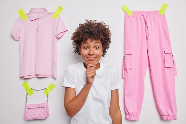 Woman with afro hair holds chin looks happily at camera wears casual t shirt poses agaist white with plastered items of clothes thinks about donation cleans out wardrobe