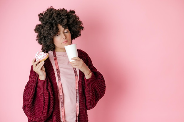 アフロヘアスタイルの女性はドーナツを持って、手に紙コップでコーヒーやお茶の温かい飲み物を楽しんでいます。測定用の巻尺が首に掛かっています。カラーコントロールのコンセプト、スマートダイエット、ピンクの背景
