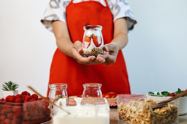 Foto donna con grembiule che tiene il parfait di fragole nel bicchiere con gli ingredienti per farlo sul tavolo