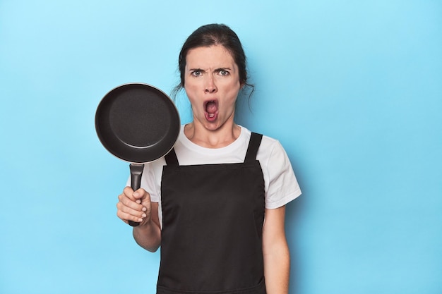 Woman with apron and pan on blue background screaming very angry and aggressive