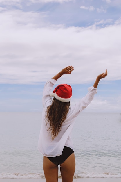 Woman with arms outstretched jumping at beach rear view of\
female is wearing santa hat and bikini carefree tourist is enjoying\
christmas vacation on sunny day