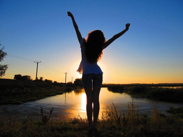 Photo woman with arms raised standing at lakeshore against sky during sunset