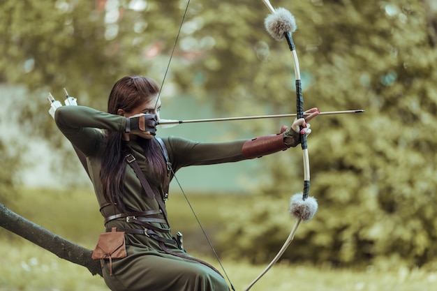 Woman with arrows and bow sits on a fallen tree and takes aim
at a target