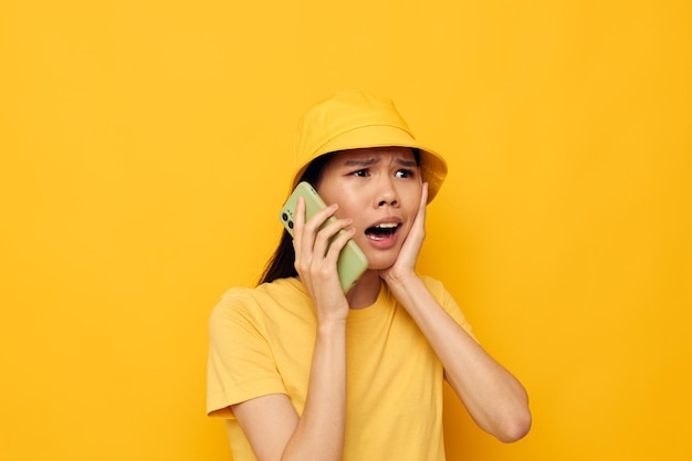 Woman with Asian appearance in a yellow hat and Tshirt talking on the phone yellow background unaltered